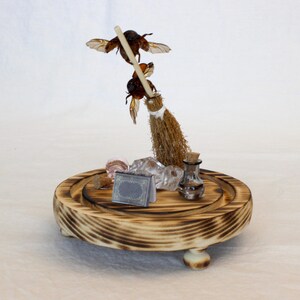 May include: A whimsical wooden display featuring a miniature broom with two insect figures. The display includes crystals, a small bottle, and a tiny book on a round, wooden base with a burnt wood finish.