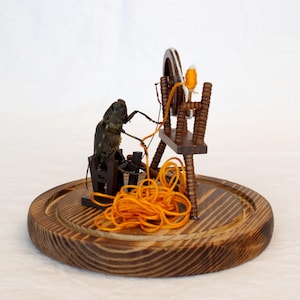 May include: A whimsical wooden sculpture featuring a large insect figure operating a miniature spinning wheel. The insect is positioned near the wheel, with orange yarn and a wooden base. The sculpture is set against a white background.