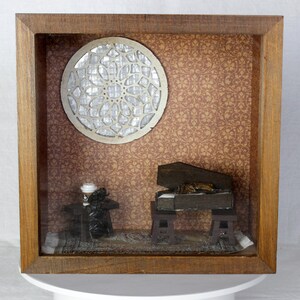 May include: A miniature diorama in a wooden frame featuring a coffin, a small table with a coffee cup, and a decorative window. The background is patterned with a brown floral design. The coffin is open, revealing its contents.