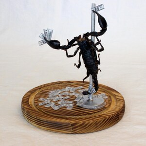 May include: A black scorpion sculpture with outstretched claws, holding and surrounded by miniature dollar bills. The scorpion is mounted on a silver pole, which is attached to a round, wooden base.