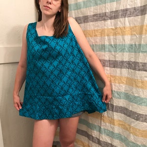 May include: A woman wearing a sleeveless, turquoise blue dress with a black geometric pattern. The dress has a square neckline and a loose, flowing fit.