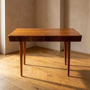 May include: A rectangular wooden table with a smooth, medium-brown finish and a visible grain pattern. The table features a flat top surface with a central seam and four tapered, cylindrical legs that extend outwards from the corners.