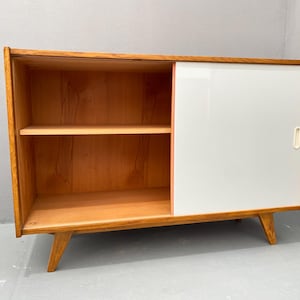 Peut inclure: Un buffet en bois de style moderne du milieu du si&egrave;cle avec une porte coulissante blanche et deux &eacute;tag&egrave;res. Le buffet est en bois clair et est sur pieds.