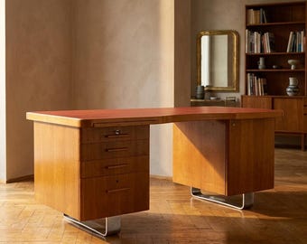 May include: A mid-century modern desk with a reddish-brown top and light wood finish. The desk features three drawers on one side and a cabinet on the other, both supported by curved metal legs. The desk is in a room with a wooden floor.