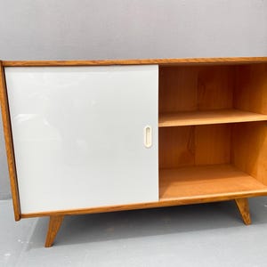 Peut inclure: Un buffet moderne du milieu du si&egrave;cle avec une porte coulissante et des &eacute;tag&egrave;res ouvertes. Le buffet est en bois avec une finition brun clair et a une porte peinte en blanc. Le buffet a deux &eacute;tag&egrave;res et repose sur quatre pieds.