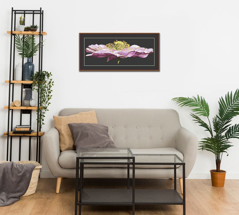 K&ouml;nnte beinhalten: Ein gerahmtes Kreuzstich-Kunstwerk, das eine gro&szlig;e, rosafarbene Blume mit gelber Mitte zeigt. Das Kunstwerk h&auml;ngt an einer wei&szlig;en Wand &uuml;ber einem grauen Sofa mit zwei Kissen.