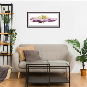 K&ouml;nnte beinhalten: Ein gerahmtes Aquarellbild einer einzelnen rosa Blume mit gelber Mitte. Das Bild h&auml;ngt an einer wei&szlig;en Wand &uuml;ber einem grauen Sofa.