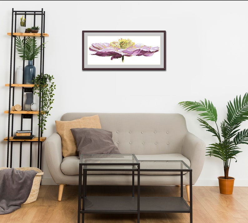 K&ouml;nnte beinhalten: Ein gerahmtes Aquarellbild einer einzelnen rosa Blume mit gelber Mitte. Das Bild h&auml;ngt an einer wei&szlig;en Wand &uuml;ber einem grauen Sofa.