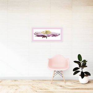 K&ouml;nnte beinhalten: Ein gerahmtes Aquarellbild einer einzelnen rosa Blume mit gelber Mitte. Das Bild h&auml;ngt an einer wei&szlig;en Wand &uuml;ber einem rosa Stuhl und einer Topfpflanze.