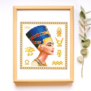 May include: A framed cross-stitch pattern of an Egyptian queen wearing a blue headdress with gold accents. The queen is surrounded by Egyptian symbols, including the Eye of Horus, the Ankh, and the Scarab beetle.