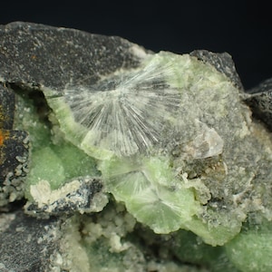Pode incluir: Imagem detalhada de uma amostra mineral com um grupo de cristais radiantes em forma de estrela. Os cristais brancos contrastam com a rocha verde e cinza, com toques de laranja e preto. A textura é áspera e natural.