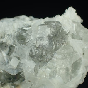 Może przedstawiać: Zbliżenie na okaz minerału z dużą, przezroczystą, szarą formacją kryształową. Kryształ otoczony jest mniejszymi, białymi strukturami krystalicznymi. Tło jest czarne, podkreślające szczegóły minerału.