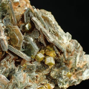 May include: Detailed image of a mineral formation. It shows a mix of green and brown, elongated, translucent crystals. There are also small, cube-shaped, yellow-green crystals. The background is dark, highlighting the rough, crystalline texture.