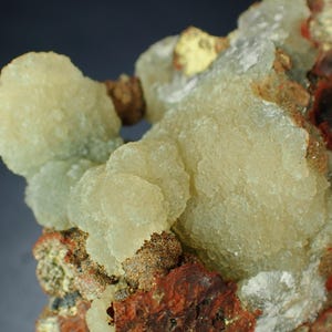 May include: Detailed view of a mineral sample with pale yellow, clustered formations. The specimen has a rough texture and is set against a backdrop of reddish-brown and tan rock.