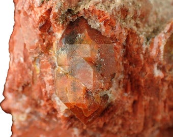 May include: Close-up of a rough, reddish-orange rock formation with a clear, faceted crystal embedded within. The crystal has a warm, amber hue and is surrounded by a matrix of coarse, textured material. The overall appearance suggests a natural mineral specimen.