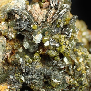 Pode incluir: Imagem detalhada de um espécime mineral. Apresenta uma mistura de pequenos cristais verdes vibrantes e cristais maiores, escuros e alongados. A superfície é áspera, com estruturas cristalinas castanhas, cinzentas e transparentes.