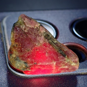 Peut inclure: Une pierre précieuse brute de couleur rouge vif, passant au rose et au vert clair. La pierre a une surface naturelle et texturée, posée sur un fond sombre.
