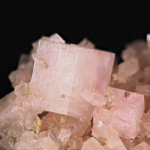 May include: A cluster of pale pink, translucent cubic mineral crystals. The specimen features sharp, geometric edges and a crystalline texture with varying levels of transparency across the interconnected, block-like formations against a black background.