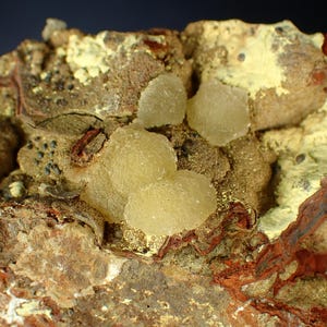 May include: A detailed view of a mineral sample. It shows clusters of pale yellow, translucent crystals on a matrix of brown, red, and yellow-green rock. The surface is rough and textured, with visible layers and colour variations.