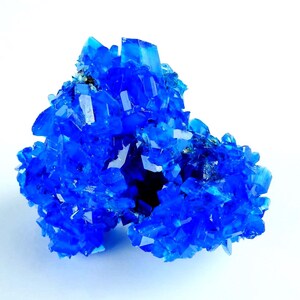 May include: A cluster of vibrant, crystalline structures in a deep, saturated blue hue. The crystals have sharp, angular facets, reflecting light and creating a sparkling effect. The mineral formation is set against a clean white background.