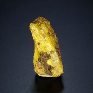 May include: A raw, natural mineral specimen with a vibrant yellow hue. The mineral has a crystalline structure with a rough, textured surface and patches of brown. The specimen is displayed against a dark background.