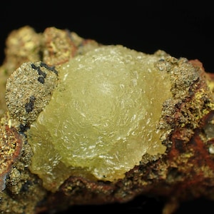 May include: A detailed image of a mineral specimen. A central, translucent, pale yellow formation with a textured surface is surrounded by a matrix of brown, gold, and reddish-brown hues, with small black inclusions. The background is black.