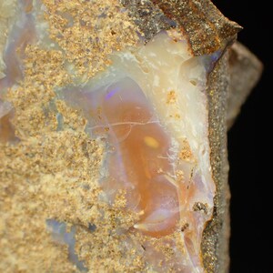 May include: A close-up of a rough, brown, and grey rock with a vein of opal. The opal is translucent and has a light pink and purple hue.