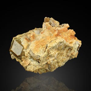 May include: A mineral specimen with a prominent, square-shaped crystal in light brown. It's set in a matrix of beige and orange minerals, offering a contrast. The texture is rough and natural. The specimen is a striking example of natural mineral formation.