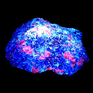 Könnte beinhalten: Ein rohes Mineralexemplar mit intensiven fluoreszierenden Eigenschaften unter ultraviolettem Licht. Der Stein weist eine raue, kristalline Textur mit einer tiefblauen Grundfarbe und verstreuten, leuchtend pinkfarbenen Einschlüssen auf, die im Dunkeln strahlen.
