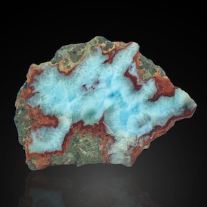 Puede incluir: Un espécimen de piedra natural con un patrón de color llamativo. La piedra presenta un centro azul claro, rodeado de blanco, con acentos rojizos y verdes. La forma general es irregular, mostrando la belleza natural y en bruto del mineral.