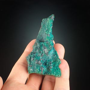 May include: A textured mineral specimen, displaying a mix of teal and dark green colours. The rough surface features small, dark inclusions, and the overall shape is irregular. The mineral is held in a hand against a dark background.