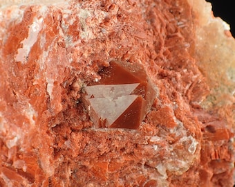 May include: Close-up of a mineral specimen featuring a cluster of reddish-brown crystals embedded in a matrix of the same color. The crystals have a geometric shape, with a lighter, almost translucent, color in the center.