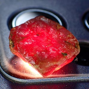 Può includere: Una gemma di rubino grezza e non tagliata, di un rosso vibrante. La gemma ha una superficie ruvida e strutturata ed è illuminata, mettendo in risalto la sua bellezza naturale. Il rubino è posto su uno sfondo scuro e strutturato.