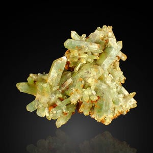 Op de afbeelding: Een cluster van lichtgroene kwartskristallen. De kristallen zijn doorschijnend en hebben een kristallijne structuur. De basis van de cluster is een mix van bruine en bruingele mineralen, tegen een donkere achtergrond.