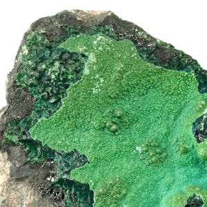 May include: A close-up of a green mineral specimen with a rough, textured surface. The mineral has a dark green colour and is surrounded by a grey, rocky matrix.