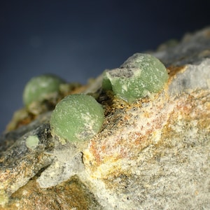 Puede incluir: Primer plano de un espécimen mineral que presenta varias formaciones pequeñas, esféricas y de color verde claro. Las formaciones están agrupadas sobre una superficie rocosa rugosa y texturizada con tonos de gris, marrón y amarillo. El fondo es un azul oscuro borroso.