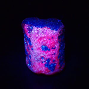 Puede incluir: Primer plano de un espécimen mineral con una vibrante combinación de colores rosa y azul. La roca tiene una superficie rugosa y texturizada con parches de azul oscuro sobre una base rosa brillante. El fondo es negro sólido.