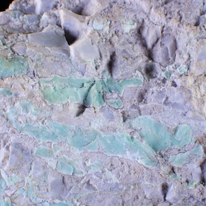 Peut inclure: Un spécimen minéral brut présentant une surface texturée avec un mélange d'inclusions gris lavande clair et vert menthe pâle. La roche affiche un aspect irrégulier et fracturé avec des profondeurs variées et des bords cristallins.