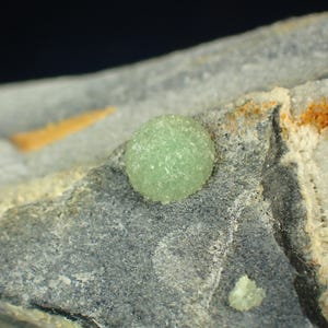 Puede incluir: Primer plano de un espécimen mineral con una pequeña formación cristalina esférica de color verde claro. El cristal se encuentra sobre una superficie rocosa gris y blanca con detalles en naranja y marrón. Fondo oscuro.