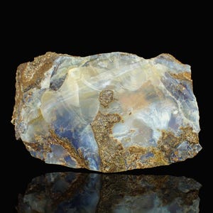 May include: A raw opal gemstone, displaying a variety of colours. The stone is translucent, featuring white, blue, and brown hues. The opal is set against a black backdrop, with a reflection visible beneath it.