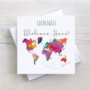 May include: A white card with a watercolour world map in shades of pink, purple, orange, and yellow. The text "HANNAH Welcome Home!" is written in black cursive script above the map. A small white paper aeroplane is flying away from the map.