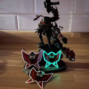 Peut inclure: Figurine détaillée de Spawn, un personnage de bande dessinée, tenant une arme à feu, avec trois autocollants du logo Spawn. La figurine est noire, rouge et grise, avec un design détaillé. Les autocollants présentent le logo Spawn en rouge et noir.