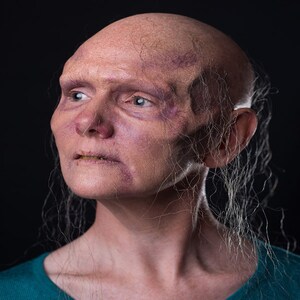 Pu&ograve; includere: Un ritratto ravvicinato di una persona con la testa rasata e un incarnato pallido. La persona ha un viso rugoso con vene visibili e guarda in alto e a destra. La persona ha capelli lunghi e bianchi che le cadono sul viso e sul collo.