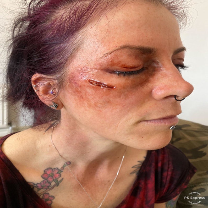 Puede incluir: Cara de una persona con maquillaje que simula una lesi&oacute;n facial. El maquillaje incluye una simulaci&oacute;n de laceraci&oacute;n y hematomas alrededor del ojo y la mejilla. La persona tiene el pelo morado y varios piercings.