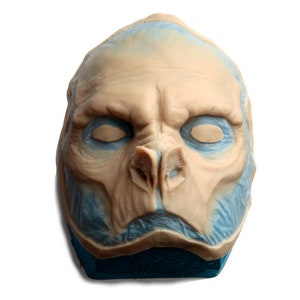 May include: A close-up of a detailed, sculpted, alien-like face mask. The mask is made of a light brown material and has a blue-grey wash applied to the edges. The mask is mounted on a blue foam block.