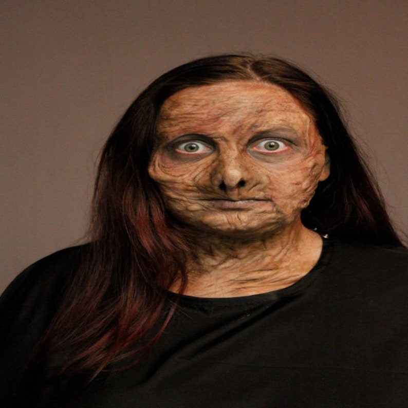 Puede incluir: Una mujer con el pelo largo y oscuro y una camisa negra tiene un maquillaje detallado, realista y envejecido en la cara. Sus ojos est&aacute;n muy abiertos y su expresi&oacute;n es seria.