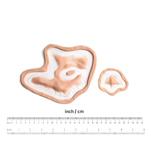 Pu&ograve; includere: Due ferite finte dall'aspetto realistico, una grande e una piccola, realizzate in silicone. Le ferite sono progettate per il trucco di effetti speciali e sono mostrate su uno sfondo bianco con un righello per la scala.
