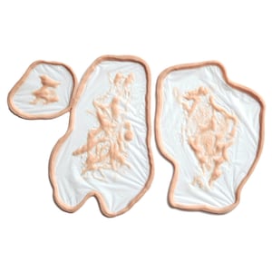 May include: Three prosthetic wound appliances. Each appliance features a flesh-toned border surrounding a white base with a textured, reddish-brown design. The shapes vary, with two larger and one smaller piece.