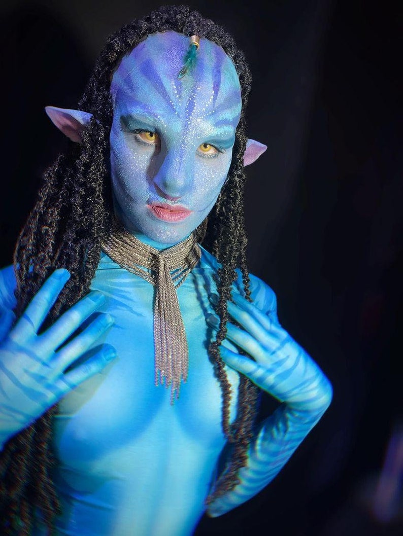 May include: A person wearing blue body paint and a silver necklace with a long tassel. They have blue and black braided hair and pointed ears. The person is posing with their hands near their chest.