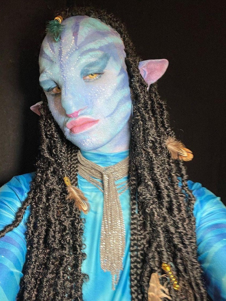 May include: A person wearing blue and white patterned clothing with long black braids and a silver chain necklace. The person has blue face paint with a design that resembles a character from the movie Avatar.